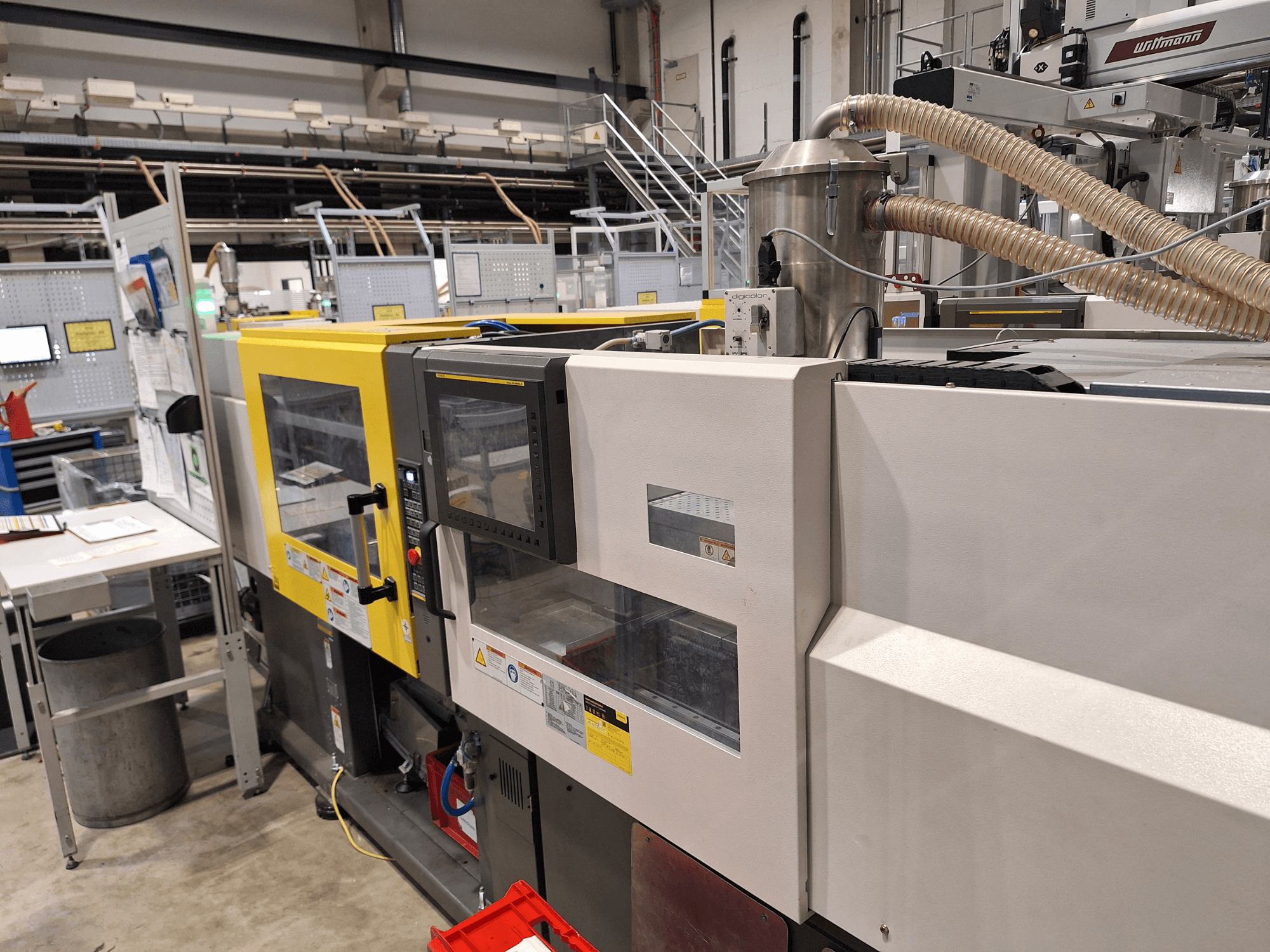 Vue de face d'une machine de moulage par injection multicolore, comprenant des sections jaunes et grises, avec panneau de commande et système d'échappement.