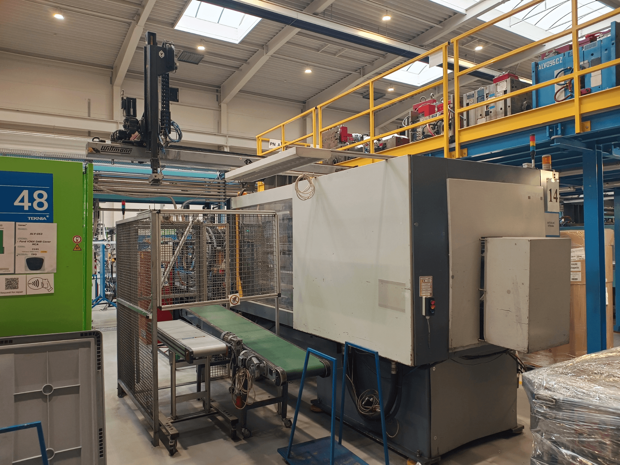 Équipement d'automatisation industrielle avec convoyeur vert, étiqueté "48", et pont roulant ; vue latérale de la machine sur le sol de l'usine.